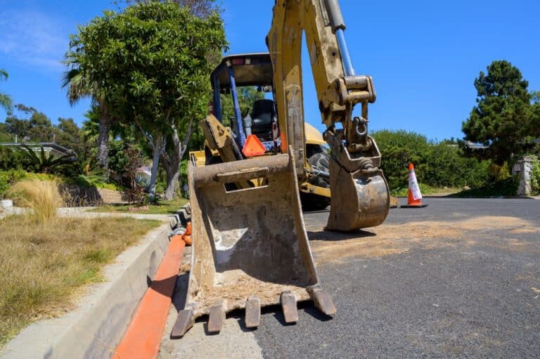 Yellow backhoe with bucket and teeth on asphalt by curb, with orange pipe and traffic cone.