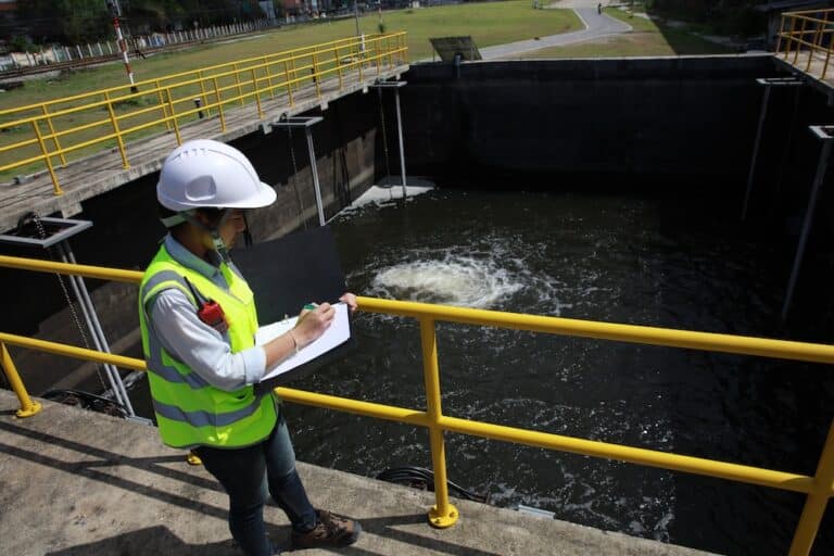 Engineer in hard hat and vest taking notes by a water treatment facility.