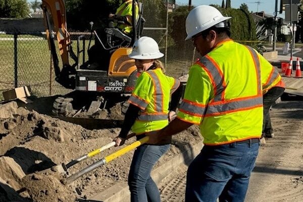 Construction workers in high-visibility vests and hard hats dig dirt with shovels near an excavator.
