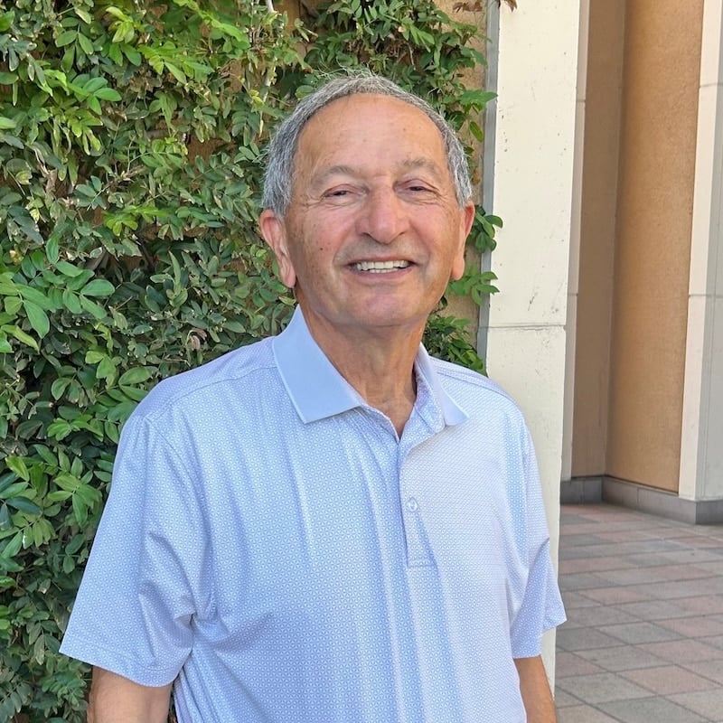 An older man with gray hair and a blue patterned polo shirt smiles warmly outdoors in front of green foliage and a building.